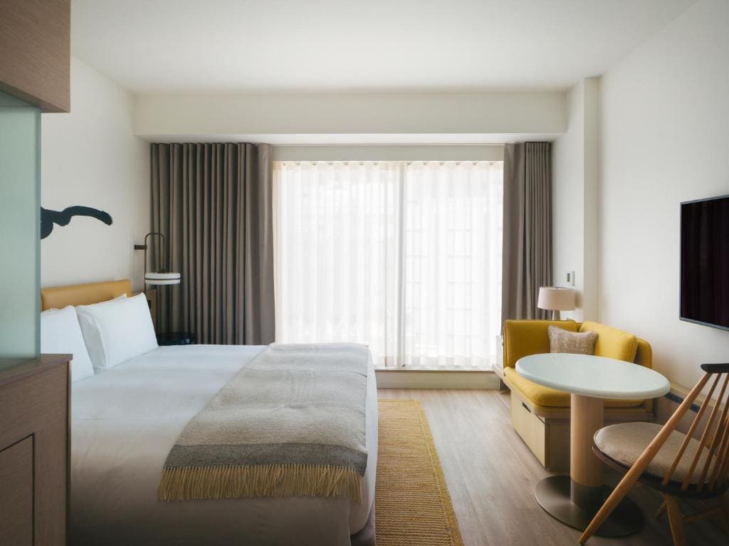 A modern hotel room with a large bed, bedside lamp, a small round table with two chairs, floor-to-ceiling window with sheer curtains, and a TV mounted on the wall. Soft neutral tones create a cozy atmosphere.