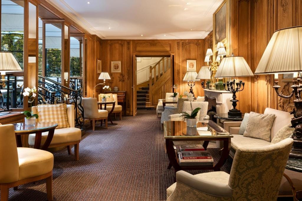 Elegant lounge with wooden-paneled walls, armchairs, sofas, and glass coffee tables with lamps. Large windows on the left let in natural light. A staircase and more seating are visible in the background.
