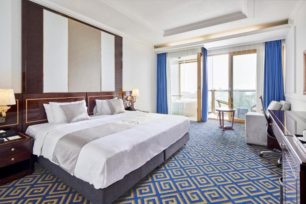 A modern hotel room with a large bed, blue geometric carpet, armchair, desk, TV, and balcony with glass doors and blue curtains, letting in natural light. The decor is neutral with wood accents.