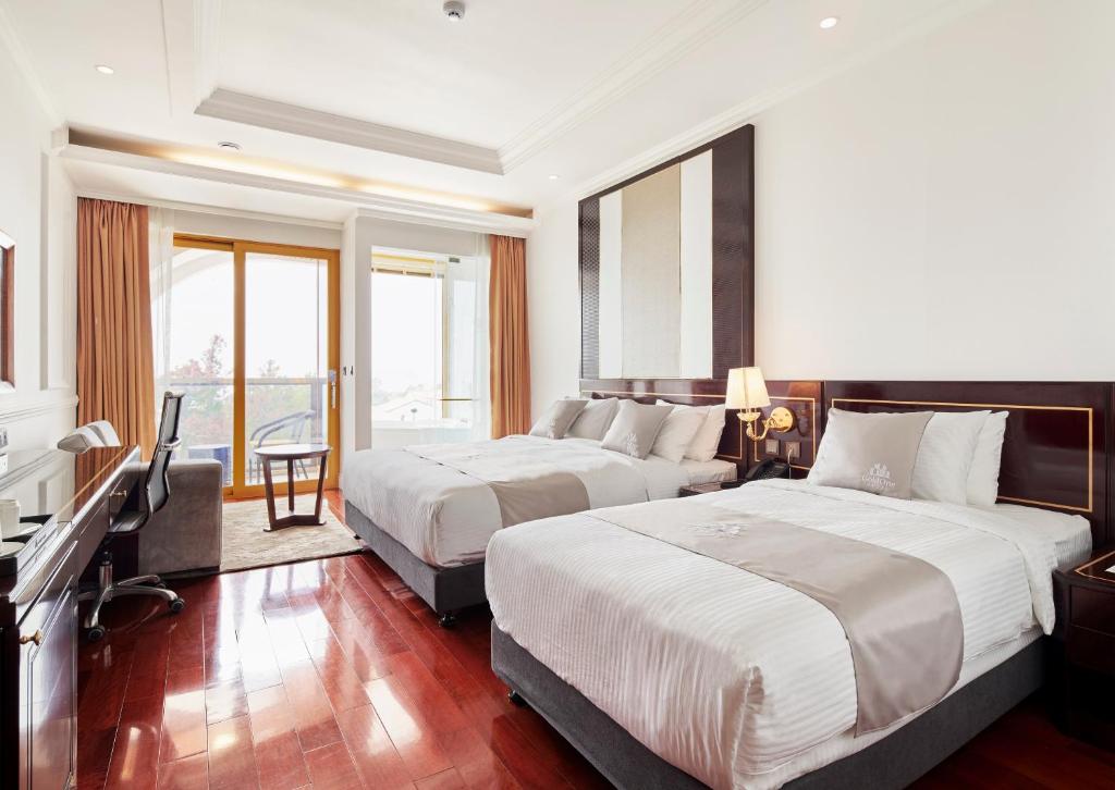 A bright hotel room with two large beds, white bedding, a desk with a chair, a sofa near a glass door leading to a balcony, and wooden floors.