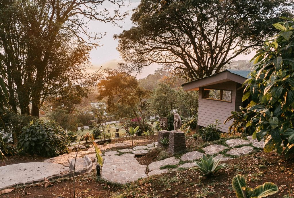 A peaceful garden scene at sunset shows a small cabin, stone pathway, lush greenery, and trees. A statue of a monkey and a sign with the number 28 are near the path. Warm sunlight filters through the trees.