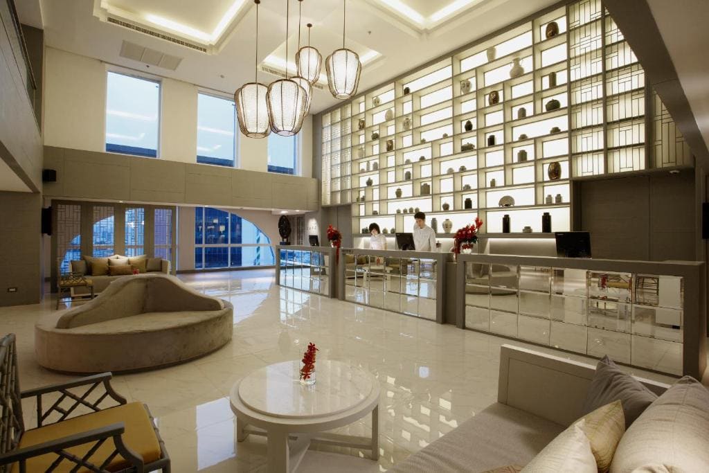 A modern hotel lobby with high ceilings, large windows, pendant lights, and a polished floor. There is stylish seating, a round table, and a backlit wall with decorative shelves behind a reception desk.