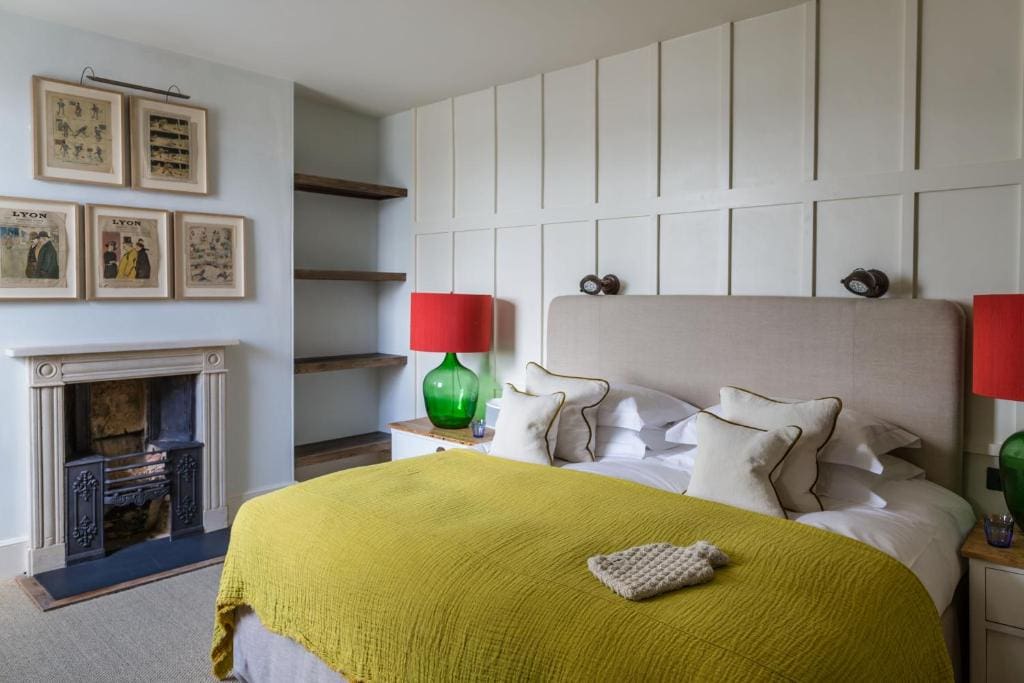 A cozy bedroom with a large bed covered by a green blanket, white pillows, two bedside tables with green lamps and red shades, a fireplace, shelves, and framed art hanging on a white paneled wall.