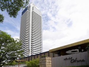 A tall, modern high-rise building stands next to a lower structure with a sign reading Hotel Okura. Trees and greenery frame the foreground under a partly cloudy sky.
