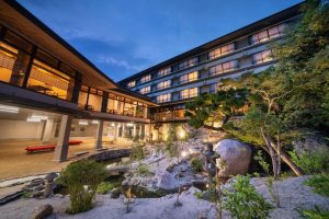 A modern Japanese-style ryokan with large windows overlooking a beautifully landscaped garden with rocks, trees, and a small pond at dusk, warmly lit from inside.