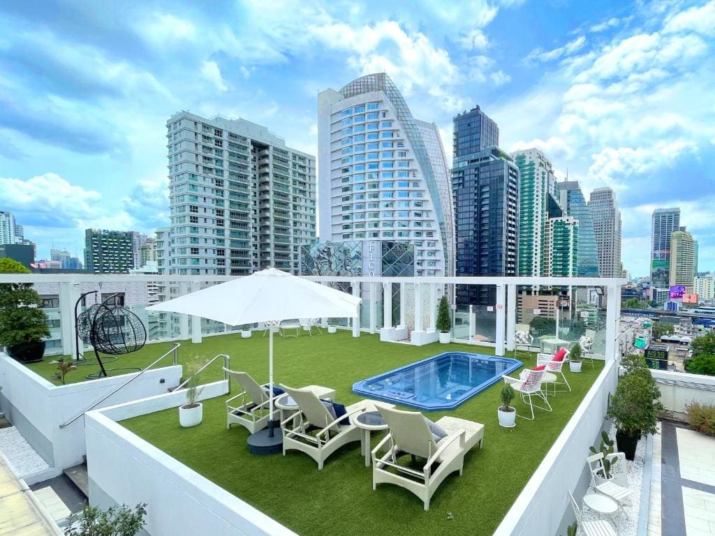 A modern rooftop terrace with artificial grass, lounge chairs, a swimming pool, umbrellas, and city skyscrapers in the background under a partly cloudy sky.
