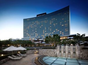 Exterior view of the Grand Hyatt hotel, a modern glass building with many lit windows, surrounded by trees. In the foreground are a small pool, stone walls, umbrellas, and fountains under a clear blue sky.