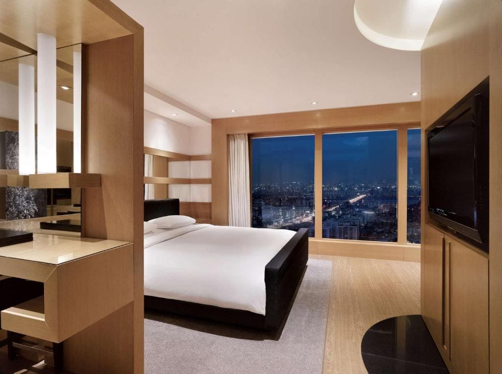 Modern hotel room with a large bed, sleek wooden furniture, a wall-mounted TV, and floor-to-ceiling windows showing a cityscape at night. Warm lighting creates a cozy and inviting atmosphere.