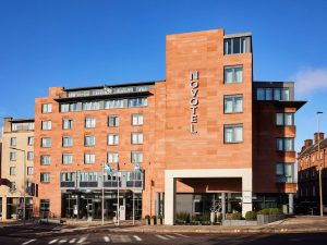 Modern multi-story hotel building with a light reddish-brown brick facade, large windows, and Novotel signage displayed vertically. Three flagpoles stand in front, and a clear blue sky is in the background.