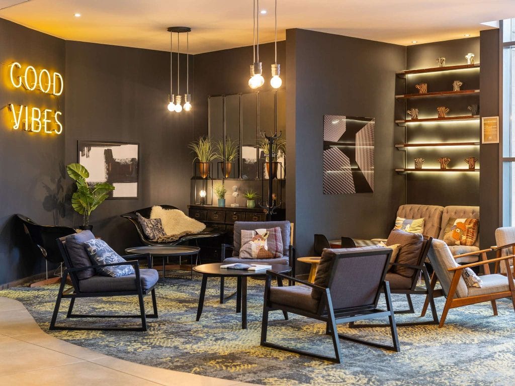 Modern lounge with dark walls, eclectic chairs, round tables, and a GOOD VIBES neon sign. Shelves with decor, plants, artwork, and warm lighting create a cozy, inviting atmosphere.