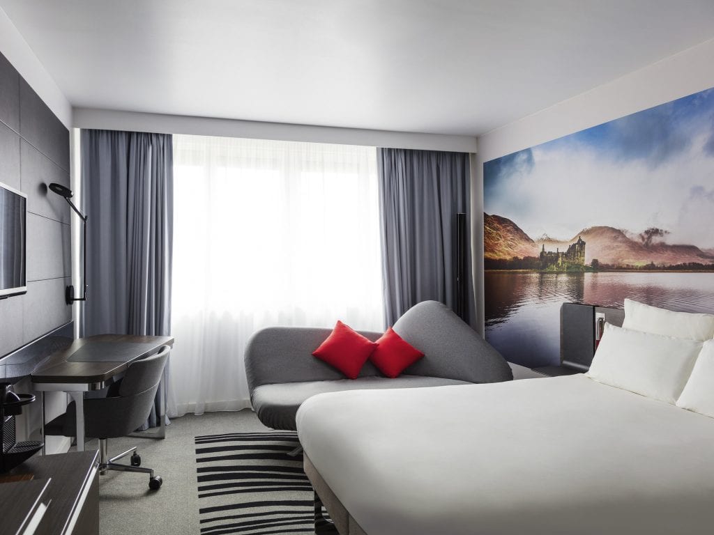 A modern hotel room with a large bed, a gray chaise lounge with red pillows, a work desk, and a wall mural of a lakeside castle and mountains. Light streams in through a large window with sheer and gray curtains.
