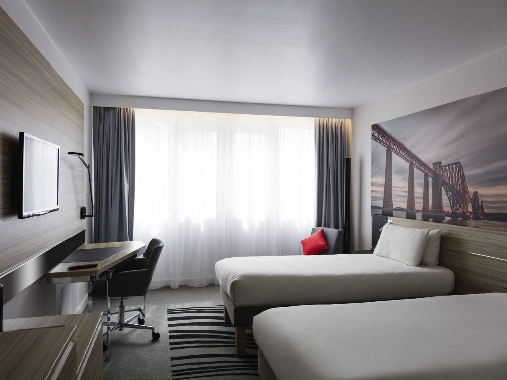 Modern hotel room with two single beds, a large window with sheer curtains, a desk and chair, a wall-mounted TV, and a wall art photo of a bridge. The room has neutral tones and a black, white, and gray rug.