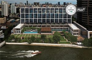 A modern, multi-story riverside hotel with large glass windows, a pool, and green gardens, set in an urban cityscape. A boat travels along the river in the foreground. An SHA Plus+ certification badge appears in the top right corner.