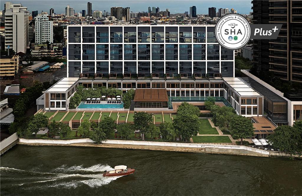 A modern, multi-story riverside hotel with large glass windows, a pool, and green gardens, set in an urban cityscape. A boat travels along the river in the foreground. An SHA Plus+ certification badge appears in the top right corner.