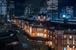 A brightly lit, historic red-brick building stands out at night among modern skyscrapers in a cityscape. People walk nearby, and the city skyline glows with lights.
