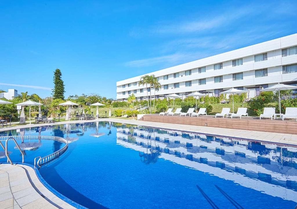 A large outdoor swimming pool with lounge chairs and umbrellas beside a modern white hotel building, surrounded by greenery and palm trees under a clear blue sky.