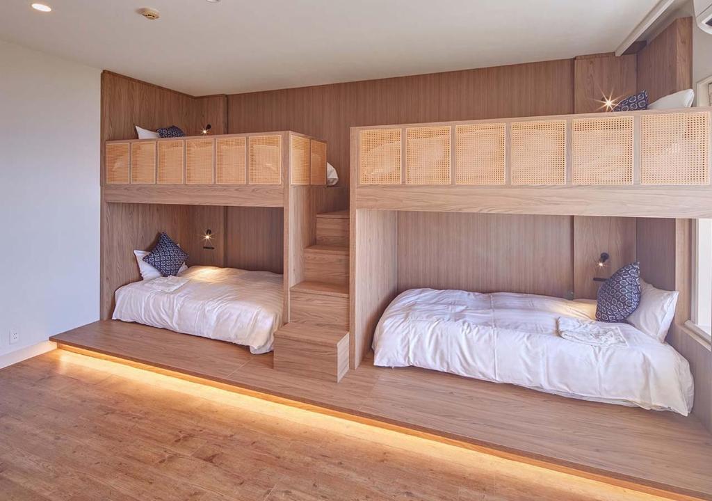 A cozy bedroom features two sets of wooden bunk beds with woven panel headboards, white bedding, patterned pillows, built-in stairs, reading lights, and warm accent lighting under the lower bunks.