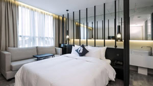 Modern hotel room with a large bed, white bedding, a gray sofa, a small table, and floor-to-ceiling window with sheer curtains. Black vertical bars separate the bed from a bathroom sink area.