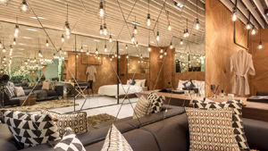 Modern hotel suite with geometric mirrors, hanging light bulbs, and wood panel walls. The room features a comfortable sofa with patterned pillows, a bed, and a cozy, stylish decor. Reflections create a spacious look.