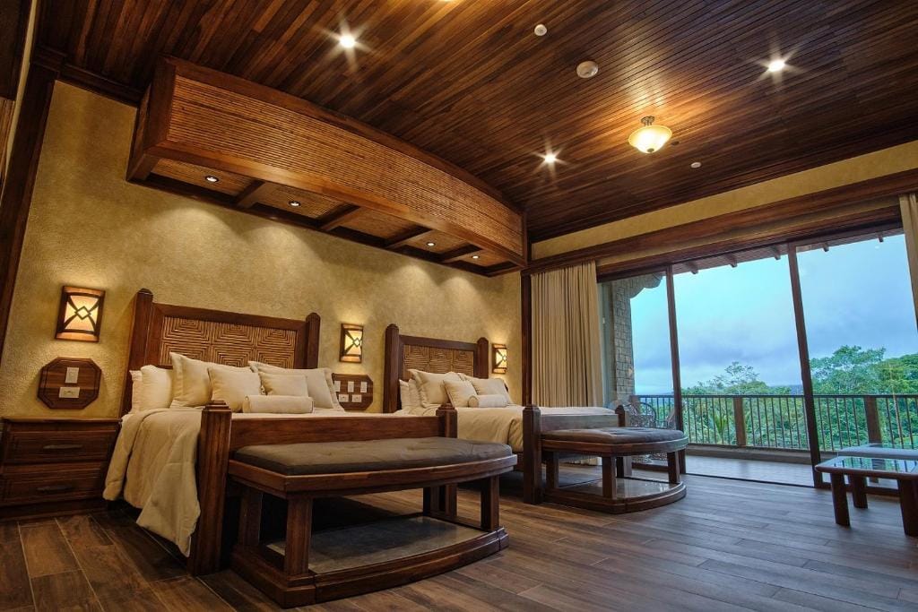 Spacious hotel room with two large beds, wooden furniture, benches at the foot of each bed, warm lighting, wooden ceiling, and a large window overlooking a green landscape.