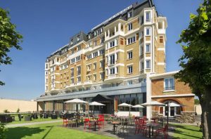 A large, multi-story brick hotel with white trim and balconies. In front, there is a patio area with red and black chairs, tables, umbrellas, and green grass. Trees frame the image under a clear blue sky.