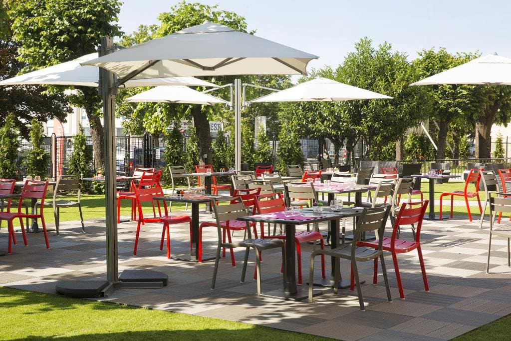 Outdoor patio seating area with red and gray chairs around tables, set on a tiled surface. Large white umbrellas provide shade, and green trees and grass surround the area on a sunny day.