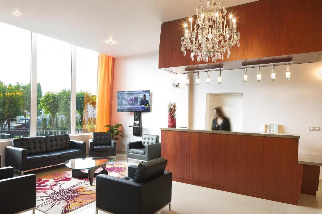 A modern hotel lobby with black leather sofas, a colorful rug, a wooden reception desk, a crystal chandelier, and large windows letting in natural light. A person stands behind the desk, with a TV mounted on the wall.