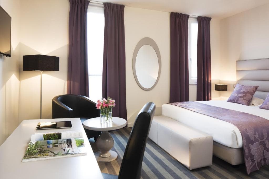 Modern hotel room with a large bed, purple accents, two tall windows with purple curtains, a round mirror, a small table with a flower vase, a desk with a magazine, and two black chairs.