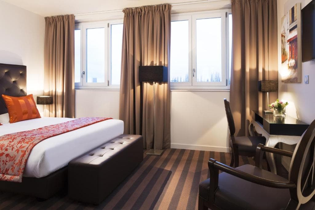 Modern hotel room with a large bed, brown and orange bedding, two brown benches, striped carpet, a desk with a chair, a lamp, and three large windows with brown curtains letting in natural light.