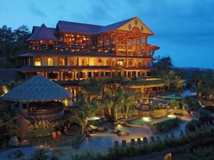 A large, warmly lit, multi-story resort with wooden architecture, surrounded by palm trees and lush gardens, overlooks multiple pools at dusk under a cloudy blue sky.