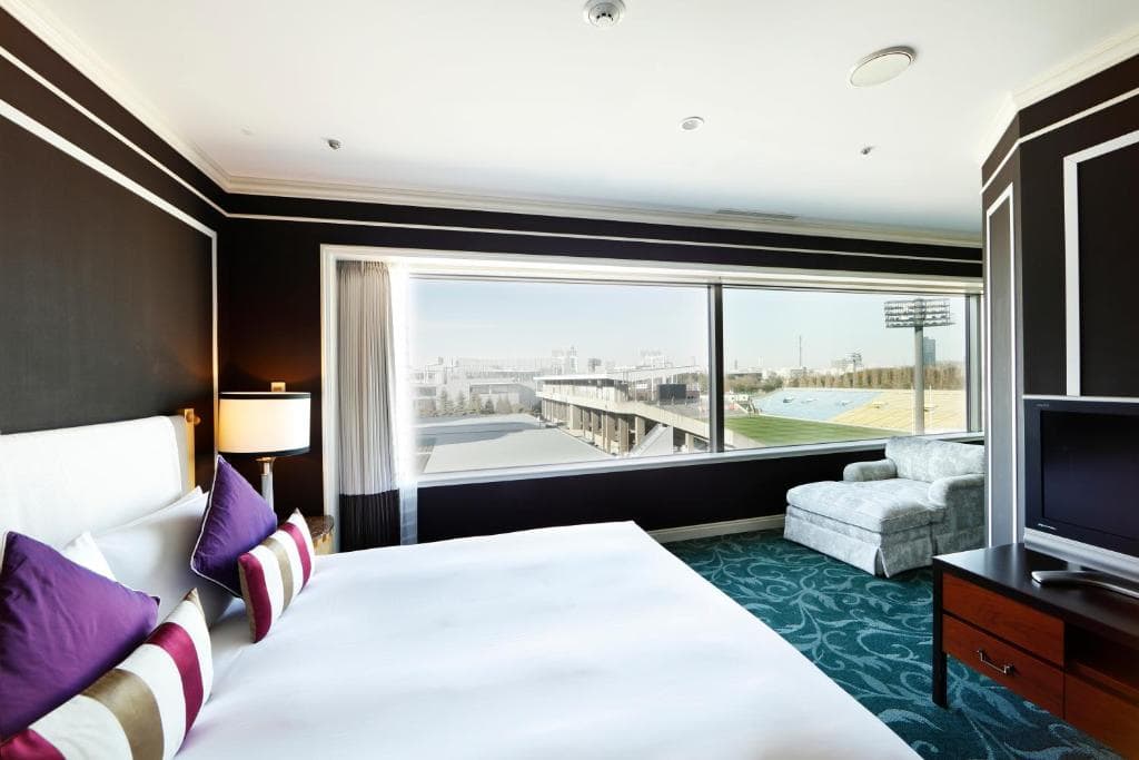 Modern hotel room with a large bed, striped pillows, a TV, a white chaise lounge, and a wide window offering a city view. The walls are dark with white trim, and natural light fills the space.