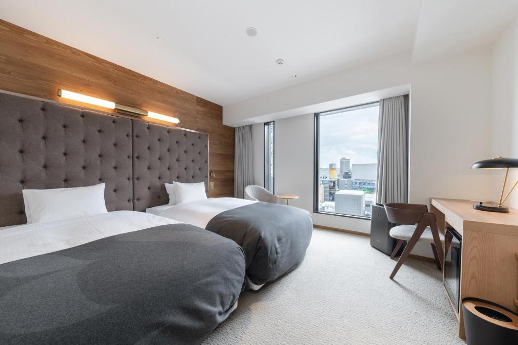Modern hotel room with two twin beds, a padded headboard, wooden accent wall, large window with city view, desk with chair, lamp, and neutral-toned decor.