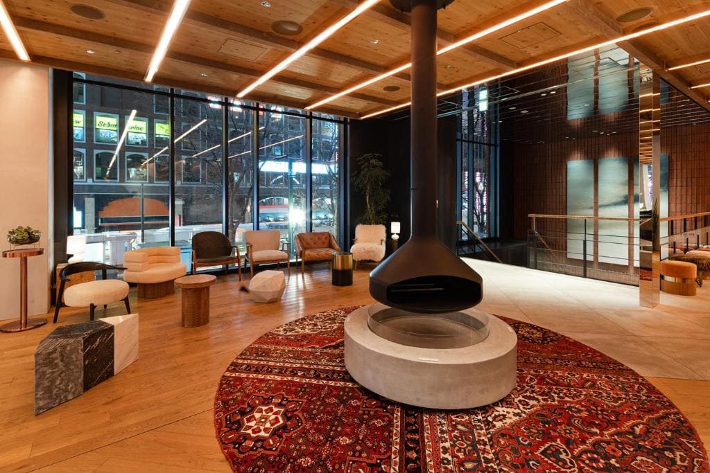 A modern lounge with a central suspended black fireplace, circular red patterned rug, and assorted chairs and tables. Large floor-to-ceiling windows reveal city lights outside. The ceiling and floor are wood paneled.