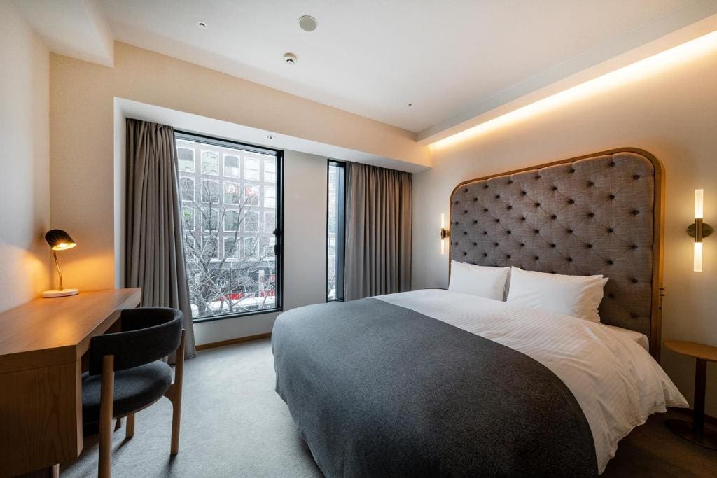 Modern hotel room with a large bed, grey upholstered headboard, desk with chair and lamp, nightstand, and large window with curtains overlooking trees and a building outside.
