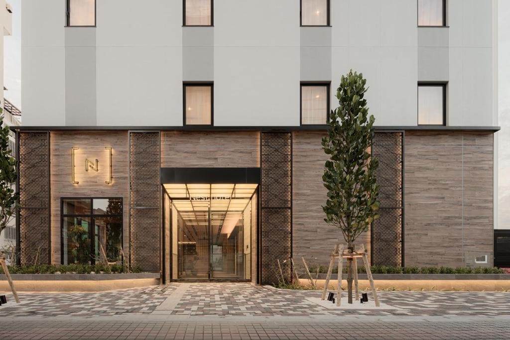 Modern building exterior with a glass entrance, large rectangular windows, and a lit sign featuring the letter N. Tall trees and landscaped planters line the walkway leading to the entrance.