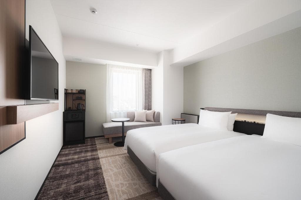 Modern hotel room with two twin beds, a wall-mounted TV, a small table with two chairs by the window, and a cabinet with a coffee maker. The room has neutral tones and minimalistic decor.
