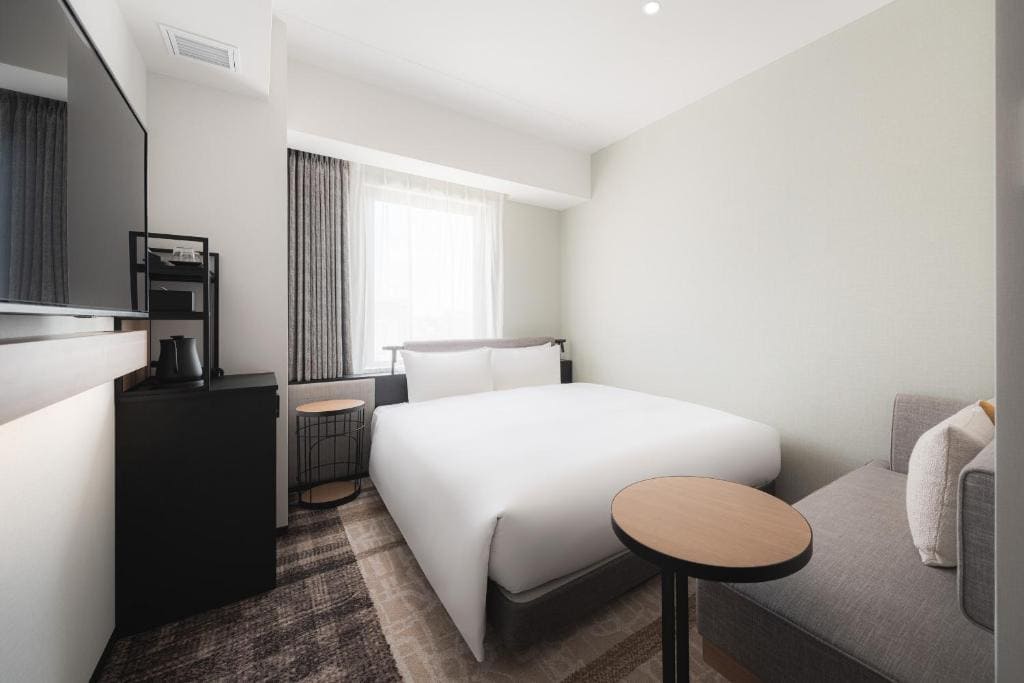 A modern hotel room with a double bed, white bedding, a gray sofa, small round tables, a wall-mounted TV, and a window with sheer curtains letting in natural light.