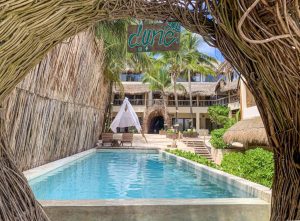 A rectangular outdoor pool is surrounded by tropical plants and thatched structures. A wooden dune sign hangs above, and there is a white canopy with lounge chairs at the far end, framed by woven branches.