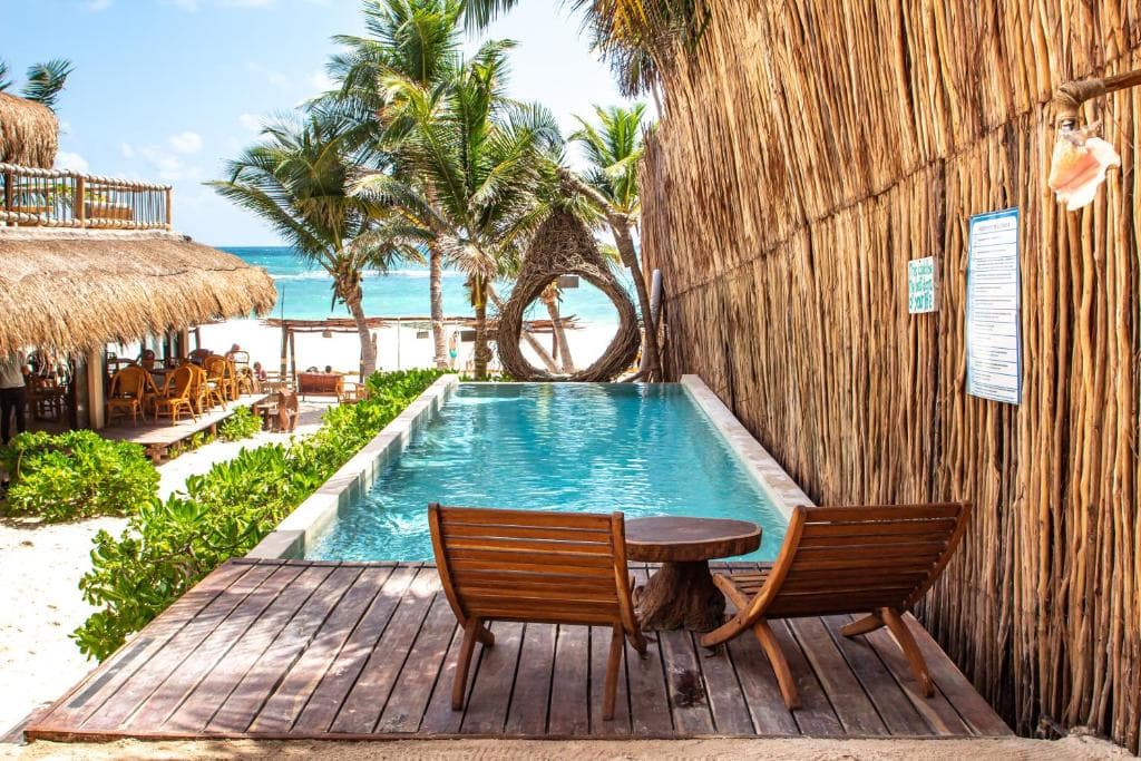 Two wooden lounge chairs and a small table sit on a deck beside a narrow pool, surrounded by tropical palms and a tall reed wall, with a sandy beach, thatched-roof structures, and ocean in the background.