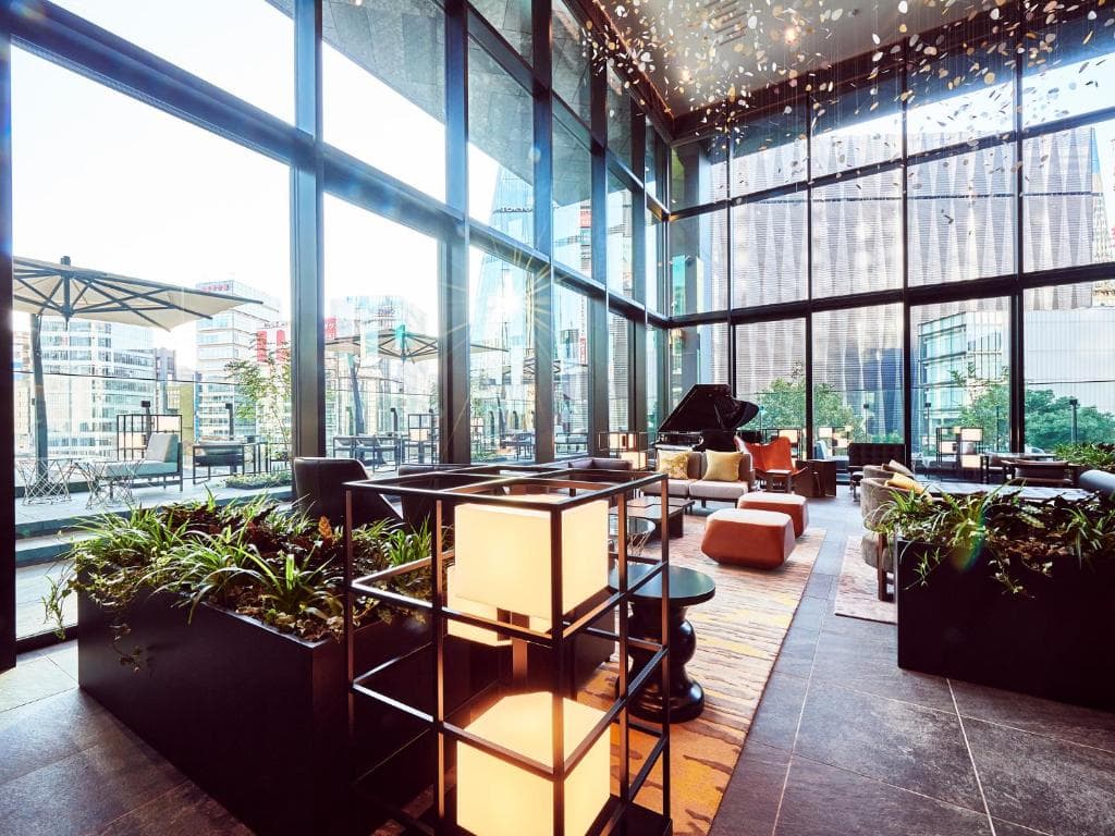 A modern, sunlit lounge with floor-to-ceiling windows, potted plants, cozy seating, a piano, and city buildings visible outside. Contemporary lights hang from the ceiling.
