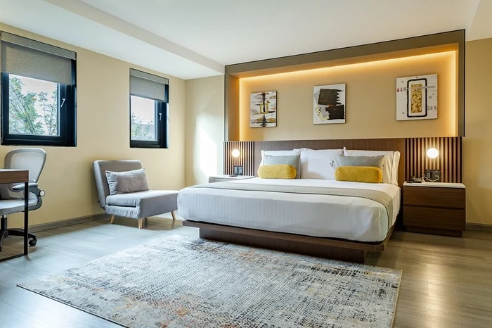 Modern hotel room with a large bed, yellow accent pillows, two bedside tables with lamps, a gray armchair, desk with a chair, and abstract art above the headboard. Light wood flooring and two windows provide natural light.