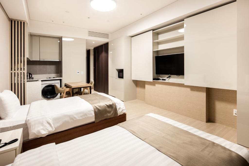 Modern hotel room with two twin beds, neutral bedding, a TV on the wall, small kitchen area with a washing machine, dining table with two chairs, and minimalist decor in beige and white tones.