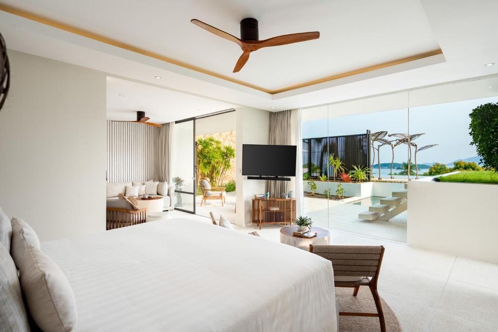 Modern, spacious hotel room with a large bed, seating area, flat-screen TV, and floor-to-ceiling windows opening to a patio with ocean and garden views. Neutral tones and natural light create a relaxing atmosphere.