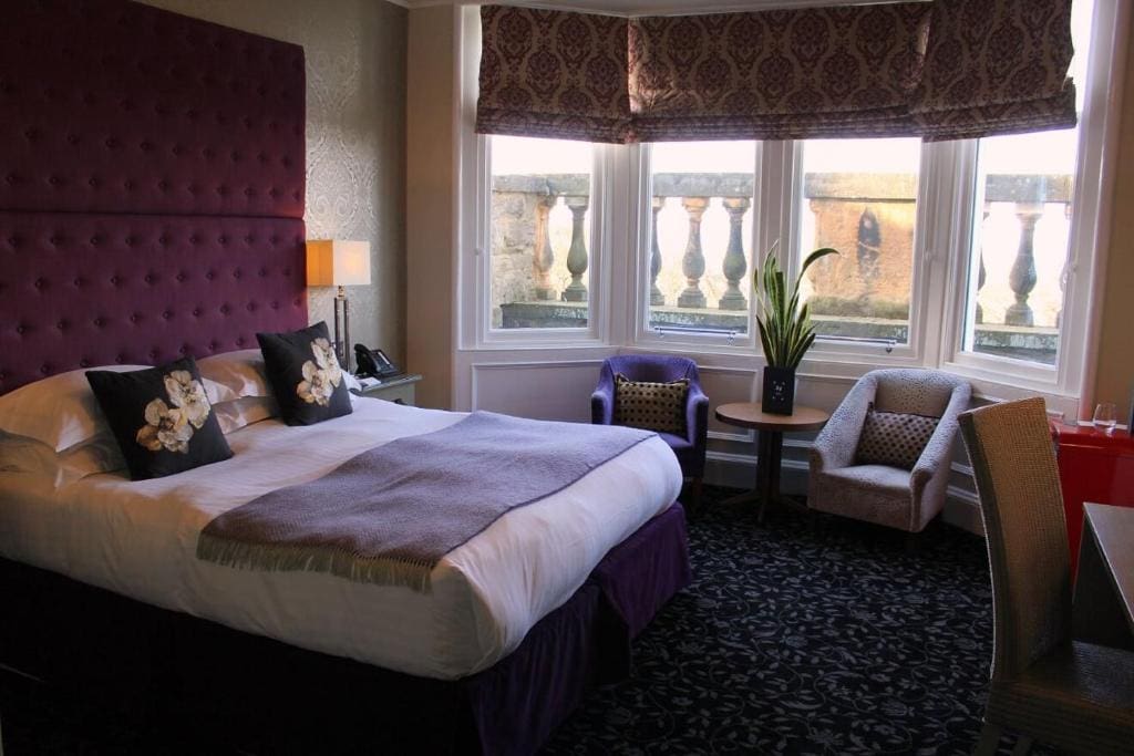 A stylish hotel room with a large bed, purple headboard, two floral pillows, a purple throw, two armchairs by a window with patterned blinds, a plant on a small table, and a desk with a chair.