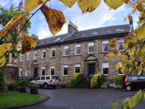 A large stone building with arched doorways and many windows, set behind trimmed bushes and parked cars. Yellow autumn leaves partly frame the scene from above.