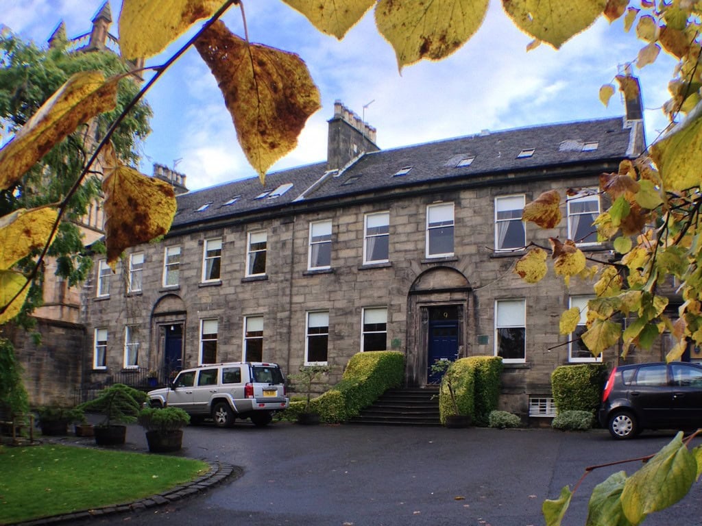 A large stone building with arched doorways and many windows, set behind trimmed bushes and parked cars. Yellow autumn leaves partly frame the scene from above.