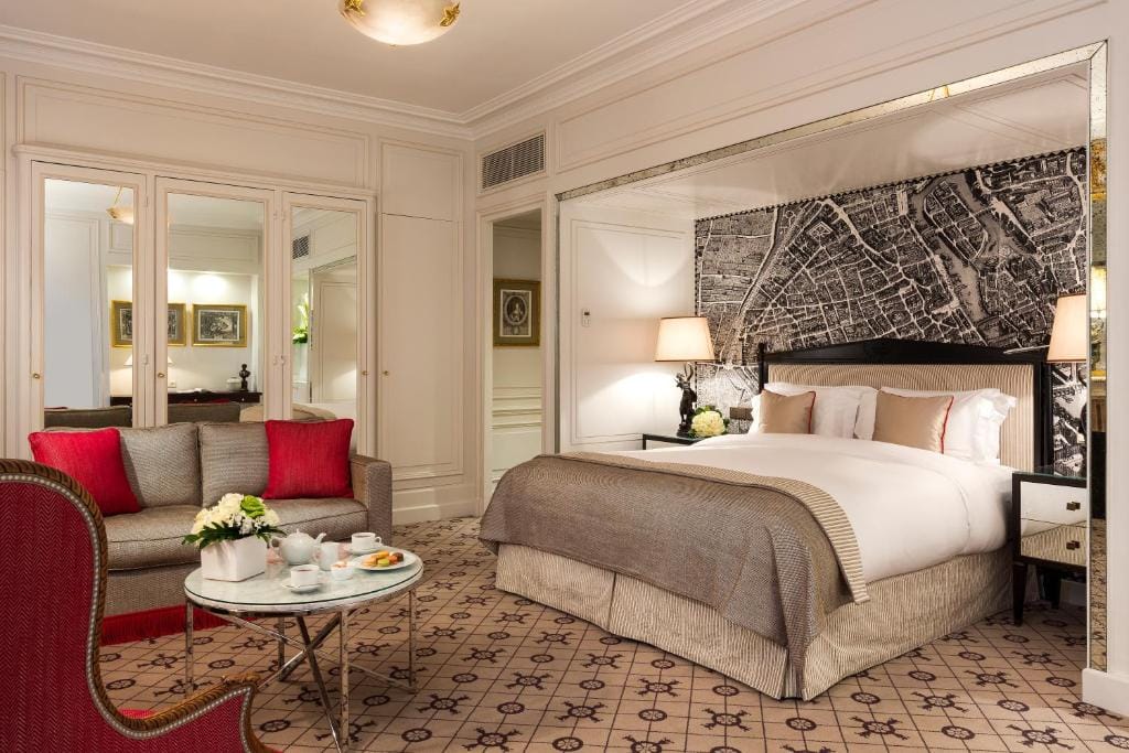 Elegant hotel room with a large bed, neutral bedding, and four pillows. There’s a sofa with red cushions, a round glass table with tea set, and mirrored wardrobes. A black and white city map decorates the wall behind the bed.