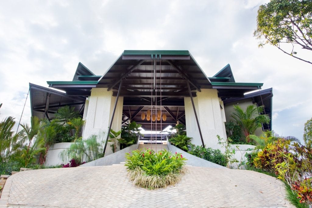 A modern building with a geometric roof, surrounded by lush green plants and landscaped gardens, viewed from a low angle on a paved walkway under a partly cloudy sky.