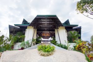 A modern building with a geometric roof, surrounded by lush green plants and landscaped gardens, viewed from a low angle on a paved walkway under a partly cloudy sky.