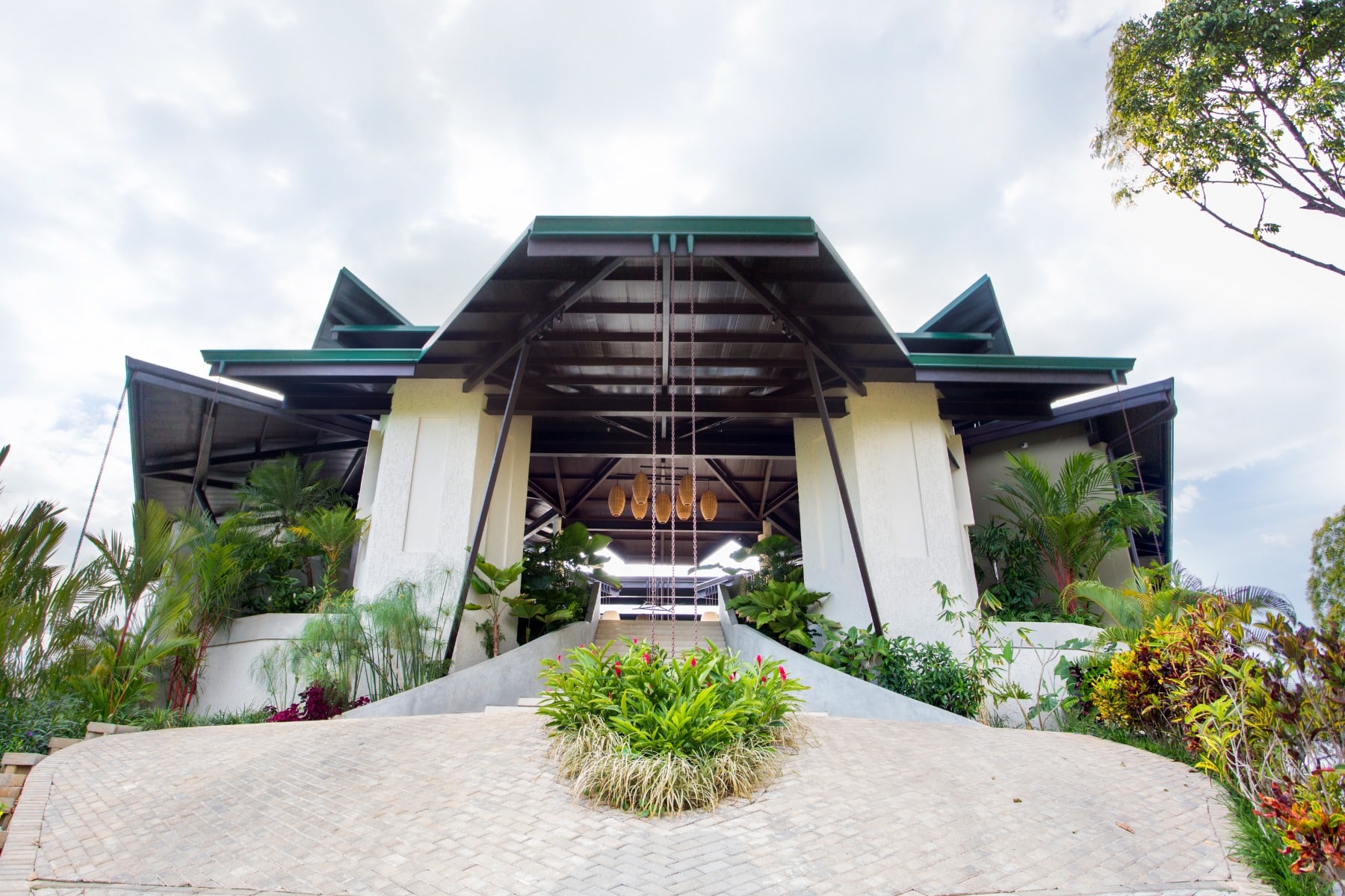 A modern building with a geometric roof, surrounded by lush green plants and landscaped gardens, viewed from a low angle on a paved walkway under a partly cloudy sky.